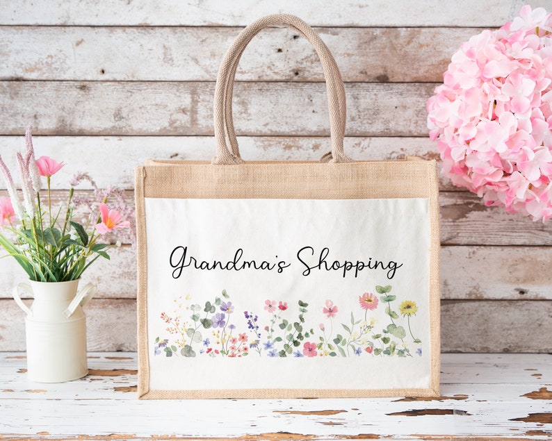 Puede incluir: Una bolsa de mano de yute natural con un panel de lona blanca con un dise&ntilde;o floral y el texto "Grandma's Shopping".