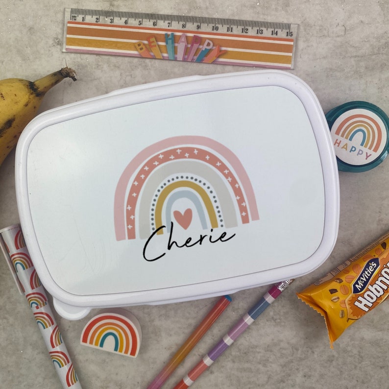 May include: White lunchbox with a rainbow design and the name "Cherie" in script. The image includes a banana, pens, a ruler, a rainbow eraser, and a snack. The lunchbox has a pink, yellow, and gray rainbow design.