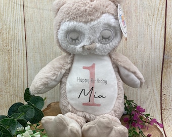May include: A plush owl toy in shades of beige and white, with closed eyes and a soft texture. The owl has a white patch on its chest with the text "Happy Birthday Mia" and the number "1" in pink. The toy is decorated with greenery and flowers.
