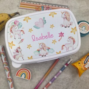 May include: White lunchbox featuring unicorn illustrations, stars, and hearts. The name "Isabelle" is written in pink. The image includes school supplies like pencils, a pen, and a rainbow eraser, creating a back-to-school theme.
