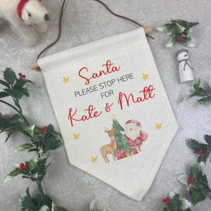 Op de afbeelding: Een crèmekleurige kerstbanner met de tekst "Santa PLEASE STOP HERE for Kate & Matt" in het rood. De banner toont illustraties van de kerstman, een rendier, een kerstboom en gouden sterren. Bovenop een houten stok en bruin koord.