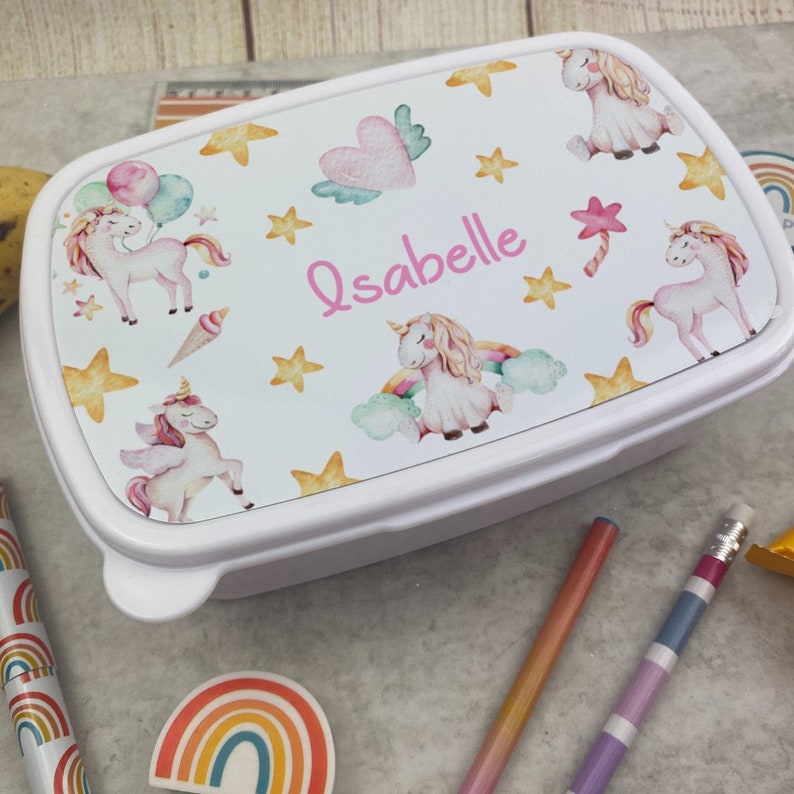 May include: White rectangular lunchbox with a lid decorated with unicorns, stars, and hearts. The lid displays the name "Isabelle" in pink script. Pencils, a rainbow sticker, and a banana are visible around the lunchbox.