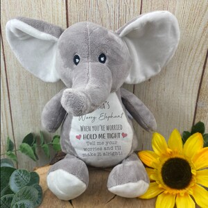 Op de afbeelding: Een grijze pluche olifant met witte accenten op de oren en voeten. De olifant heeft de tekst "Mama's Worry Elephant" en "HOLD ME TIGHT" op de borst gedrukt. Een gele zonnebloem staat op de achtergrond.