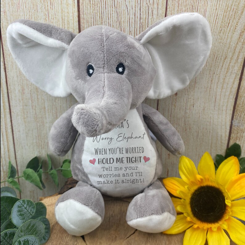 Op de afbeelding: Een grijze pluche olifant met witte accenten op de oren en voeten. De olifant heeft de tekst "Mama's Worry Elephant" en "HOLD ME TIGHT" op de borst gedrukt. Een gele zonnebloem staat op de achtergrond.