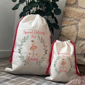 Puede incluir: Dos bolsas de regalo con cordón de color crema con cordones rojos. Las bolsas presentan un diseño de acuarela de una bailarina dentro de una corona verde con el texto "Special Delivery For Anna". Las bolsas están sobre una alfombra estampada.