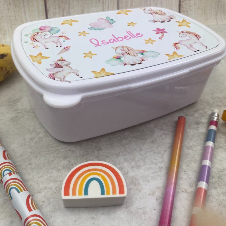 May include: White rectangular lunchbox featuring a lid adorned with unicorns, stars, and clouds in watercolour. The name "Isabelle" is written in pink script. A rainbow eraser and pencils are also visible.