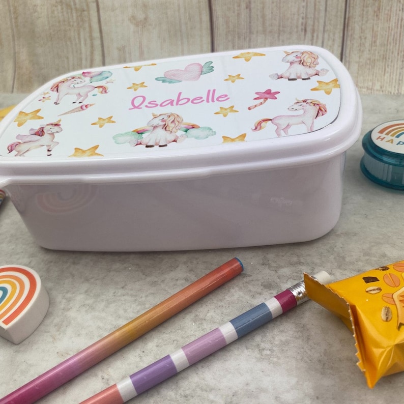 May include: White plastic lunchbox featuring a lid adorned with watercolour unicorns, stars, and hearts. The name "Isabelle" is printed in pink. Pencils, a rainbow eraser, and a snack are also visible.