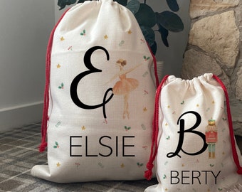 May include: Two off-white drawstring gift bags with red cords. One bag features a ballerina illustration and the name "Elsie", the other has a nutcracker illustration and the name "Berty". Both bags have festive star and holly accents.