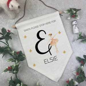 May include: A cream-colored banner with the text "SANTA PLEASE STOP HERE FOR & ELSIE". The banner features a black ampersand, gold stars, and a ballerina in a pink tutu. A wooden dowel and brown cord are at the top for hanging.