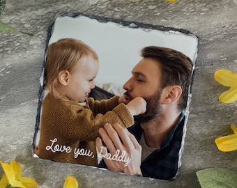 May include: A black slate coaster with a photo of a father and child. The child is holding onto the father's beard and smiling. The text "Love you, Daddy" is written in white on the coaster.