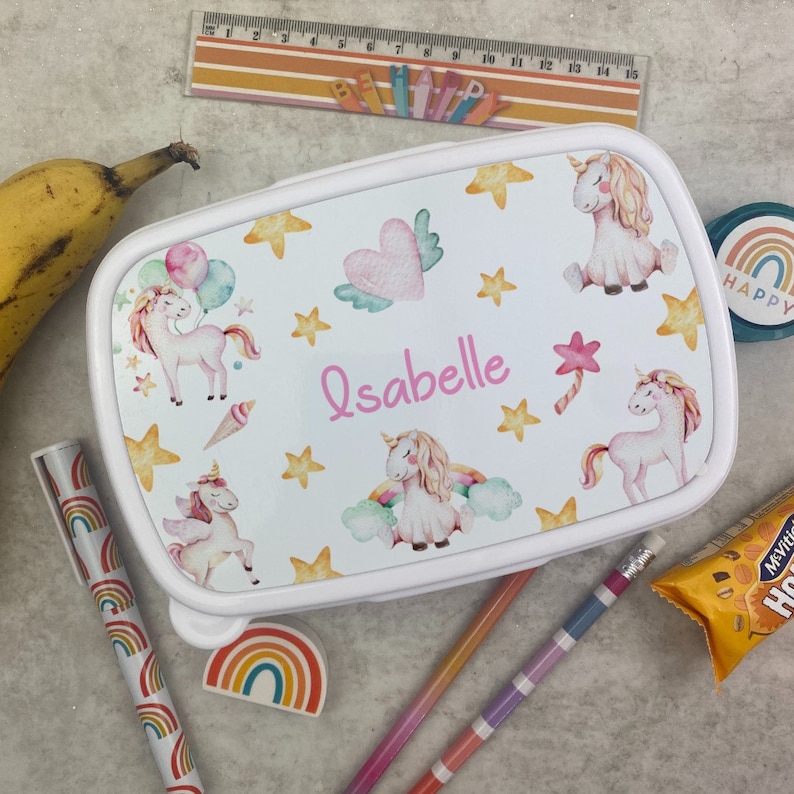 May include: A white lunchbox featuring unicorn illustrations, stars, and a heart with wings. The name "Isabelle" is written in pink. A ruler, pencils, and a rainbow-themed pen are also visible.