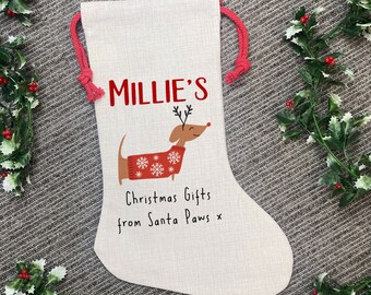 Może przedstawiać: Beżowa pończocha świąteczna z czerwonym sznurkiem. Zawiera napis "Millie's" w kolorze czerwonym, kreskówkowego jamnika w czerwonym swetrze ze śnieżynkami oraz tekst "Christmas Gifts from Santa Paws x."
