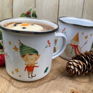 Puede incluir: Dos tazas de esmalte blanco con bordes plateados, una llena de chocolate caliente y una malvavisco de muñeco de nieve. Las tazas presentan ilustraciones festivas de un elfo y un muñeco de nieve, con estrellas doradas y acentos de acebo.