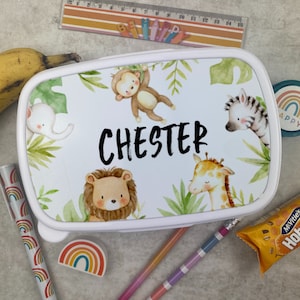 Puede incluir: Fiambrera rectangular blanca con dise&ntilde;o de animales de la jungla. La fiambrera presenta un mono, un elefante, un le&oacute;n, una cebra y una jirafa, con el nombre "CHESTER" en negro. El dise&ntilde;o incluye hojas verdes. Otros art&iacute;culos incluyen un pl&aacute;tano, l&aacute;pices y una regla.
