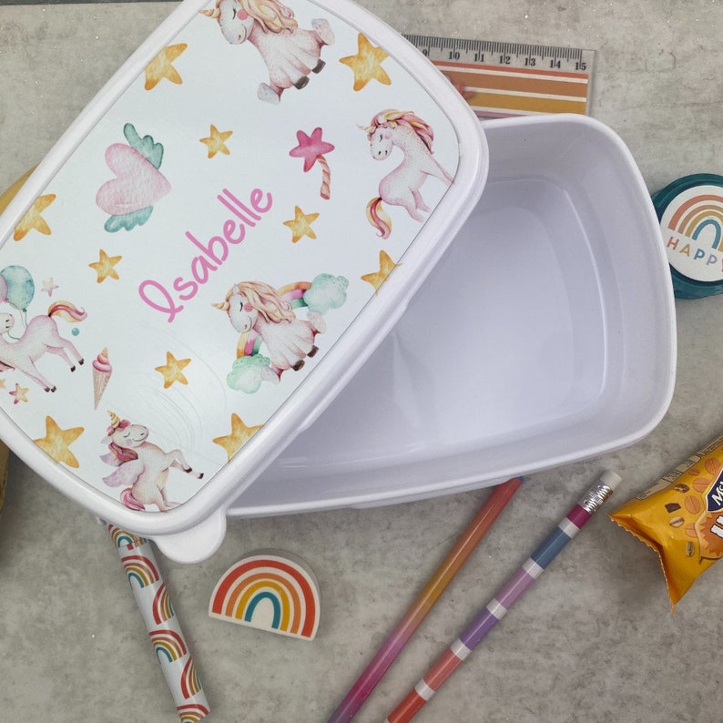 May include: White rectangular lunchbox with a lid decorated with unicorn illustrations, stars, and the name "Isabelle". The lunchbox is open, showing the empty interior. Pencils and other stationery are also visible.