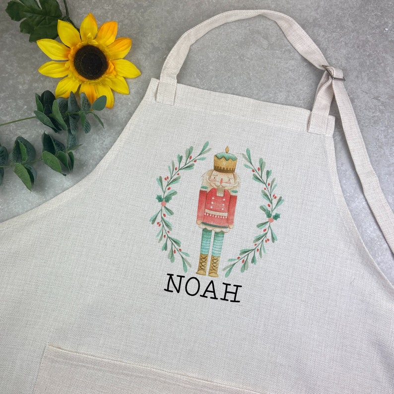 Puede incluir: Delantal blanquecino con una ilustraci&oacute;n de acuarela de un cascanueces dentro de una corona verde. El cascanueces lleva una chaqueta roja y pantalones turquesa. El nombre "NOAH" est&aacute; impreso debajo. Un girasol amarillo y hojas verdes en el fondo.