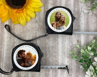 May include: Two black measuring tapes with custom photo inserts. One tape reads "DADDY LOVED BEYOND MEASURE" and "LOVE BEN X" with a photo of a father and child. The other reads "DAD NO ONE MEASURES UP TO YOU" and "LOVE MIA & MAISIE X" with a photo of two girls. A sunflower and greenery are in the background.