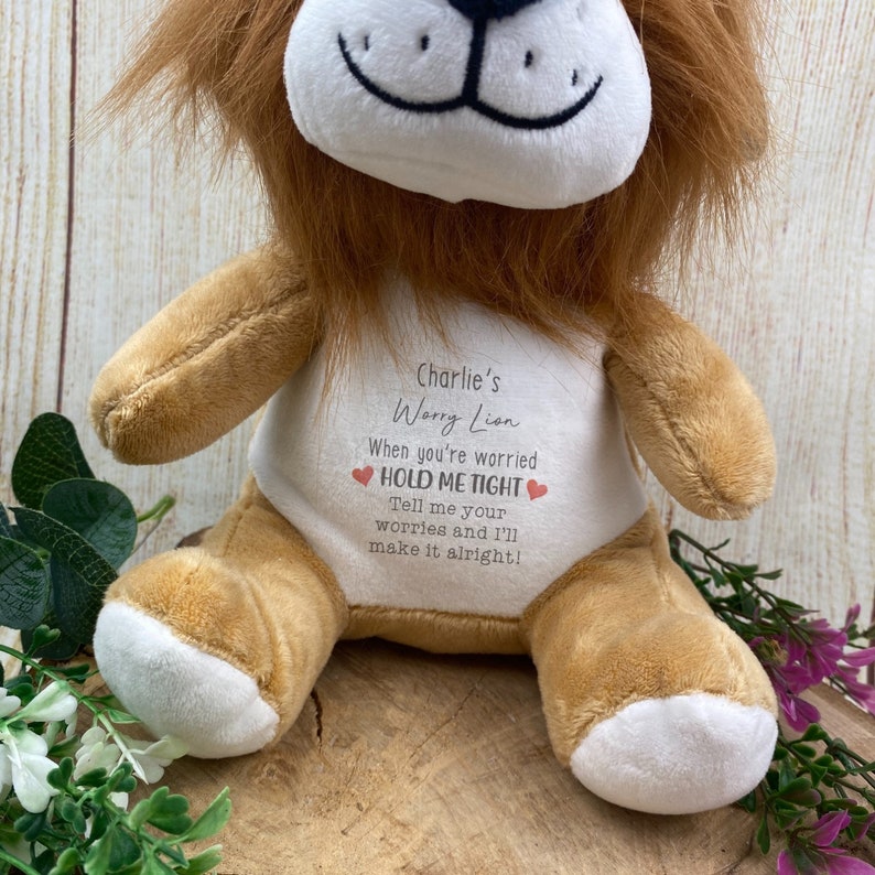 Op de afbeelding: Een pluche leeuw met een bruine manen en een bruin lichaam. De leeuw heeft een witte buik met de tekst "Charlie's Worry Lion" en de zin "HOLD ME TIGHT". Het speelgoed is ontworpen om te troosten en gerust te stellen.