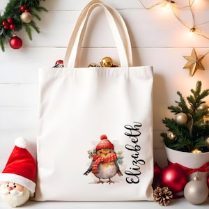 Puede incluir: Un bolso tote blanco con una ilustración de acuarela de un pájaro con gorro y bufanda rojos. El nombre "Elizabeth" está escrito en cursiva. El bolso tiene dos asas blancas y está rodeado de adornos navideños.