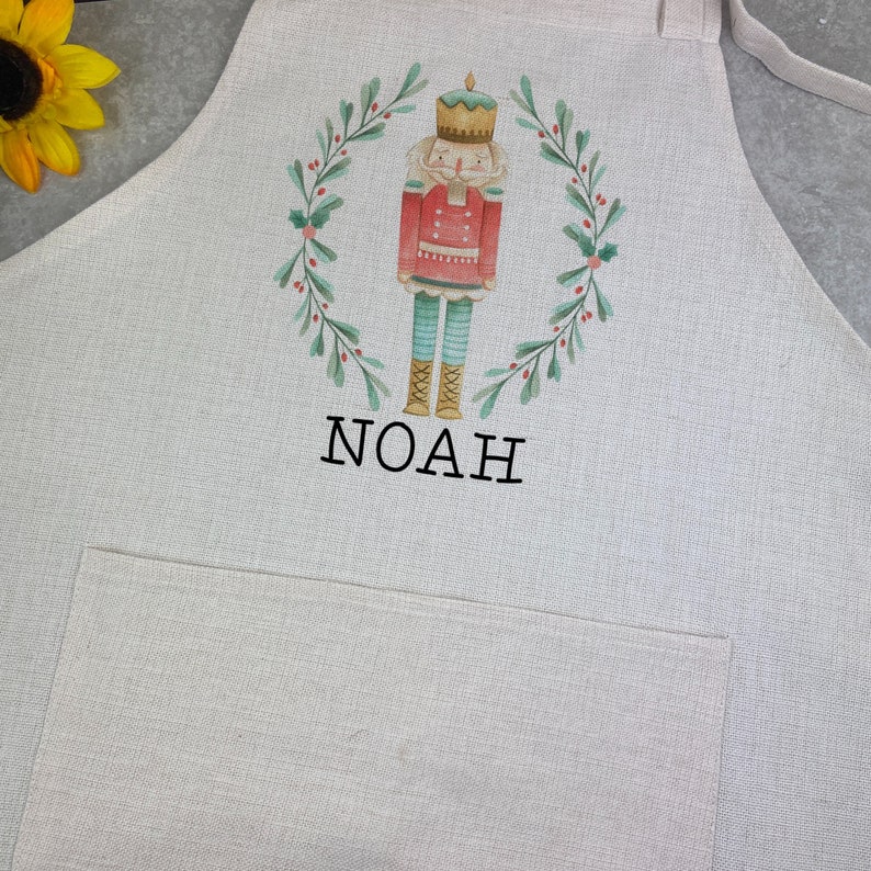 Puede incluir: Un delantal de color claro con un bolsillo y lazos. El delantal presenta una ilustraci&oacute;n de acuarela de un cascanueces dentro de una corona verde. El nombre "NOAH" est&aacute; impreso en negro debajo.