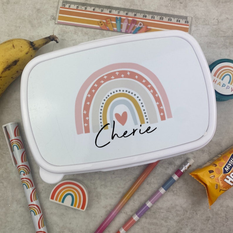 May include: White lunchbox with a rainbow design, a heart, and the name "Cherie" in black script. The lunchbox is surrounded by school supplies, including pencils, a ruler, and a banana.