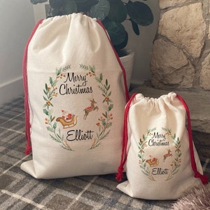 Puede incluir: Dos sacos de Santa de lona blanca con cordones de extracción rojos. El saco más grande tiene un diseño de corona verde con el texto "Merry Christmas Elliott". El saco más pequeño tiene el mismo diseño con el texto "Merry Christmas Elliott".