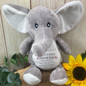 Op de afbeelding: Een grijze pluche olifant met grote oren en een slurf. De olifant heeft een witte buik met de tekst "Elephant, YOU'RE WORRIED, HOLD ME TIGHT, Tell me your worries and I'll make it alright!" Een gele zonnebloem staat op de achtergrond.