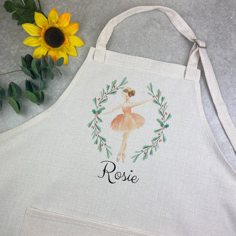 Puede incluir: Un delantal color crema con una ilustraci&oacute;n en acuarela de una bailarina con un tut&uacute; melocot&oacute;n, enmarcada por una corona verde con bayas rojas. El nombre "Rosie" est&aacute; escrito debajo en escritura negra. El delantal tiene un bolsillo y una correa ajustable para el cuello.