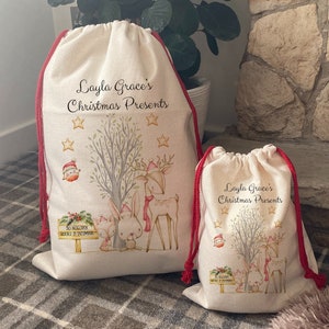 May include: Two Christmas gift bags with red drawstrings. The bags are beige with a woodland scene featuring a deer, rabbit, and bird. Text on the bags reads "Layla Grace's Christmas Presents" and "Do Not Open Before 25 December".