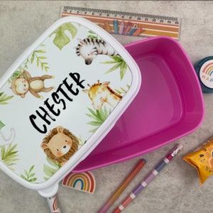 Puede incluir: Una fiambrera blanca con interior rosa, con un dise&ntilde;o de animales de la selva: cebra, mono, jirafa y le&oacute;n. El nombre "Chester" est&aacute; impreso en negro. Hay &uacute;tiles escolares alrededor.