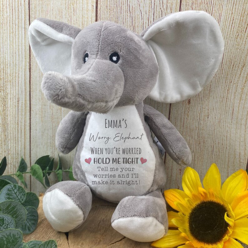 Op de afbeelding: Een grijze pluche olifant met grote oren en een slurf. De olifant heeft een witte patch op zijn borst met de tekst "EMMA'S Worry Elephant" en de zin "HOLD ME TIGHT". Een gele zonnebloem staat op de achtergrond.