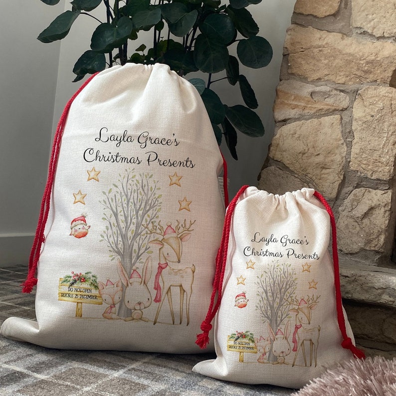 May include: Two off-white Christmas gift bags with red drawstring closures. The bags feature a watercolor illustration of woodland animals, stars, and the text "Layla Grace's Christmas Presents".