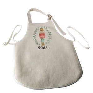 Puede incluir: Un delantal beige con una ilustraci&oacute;n en acuarela de un cascanueces dentro de una corona verde. El cascanueces es rojo, verde y dorado. El nombre "NOAH" est&aacute; impreso en negro debajo. El delantal tiene una correa para el cuello y lazos en la cintura.
