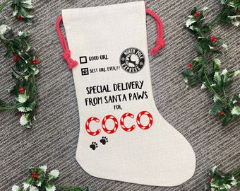 Może przedstawiać: Beżowa skarpeta świąteczna z czerwonymi akcentami i tekstem. Skarpeta zawiera napisy "GOOD GIRL" i "BEST GIRL EVER!!" z logo "North Pole Express". Imię "COCO" jest wyświetlane czerwonymi literami w kształcie laski cukrowej, z odciskami łap poniżej.