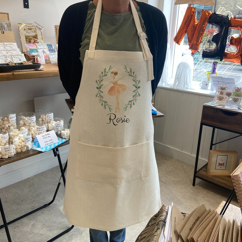 Puede incluir: Delantal beige con un dise&ntilde;o de bailarina dentro de una corona verde. El nombre "Rosie" est&aacute; escrito debajo. Tiene un bolsillo frontal grande y una correa ajustable para el cuello. El delantal es de tela de color natural.