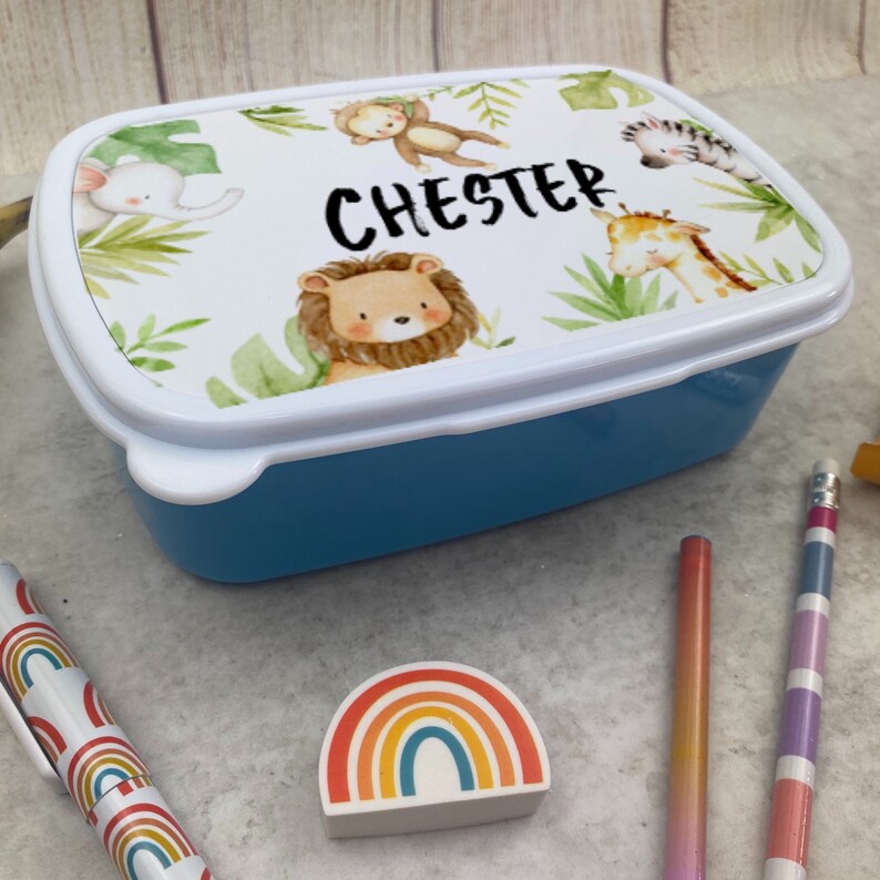 Puede incluir: Una fiambrera azul con tapa blanca decorada con animales de la selva y el nombre "CHESTER". La tapa presenta un le&oacute;n, un mono, una jirafa, un elefante y una cebra, rodeados de follaje verde. Ideal para ni&ntilde;os.