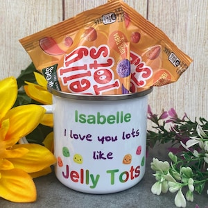 Pode incluir: Caneca de esmalte branco com uma mensagem personalizada que diz "Isabelle, eu te amo muito como Jelly Tots". A caneca está cheia de duas pequenas sacolas de doces Jelly Tots laranja e amarelos.