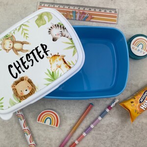 Puede incluir: Una fiambrera azul con tapa blanca con animales de la jungla y el nombre "Chester". La tapa tiene una ilustraci&oacute;n de mono, cebra, jirafa y le&oacute;n. Otros art&iacute;culos incluyen l&aacute;pices, una regla y un bocadillo.