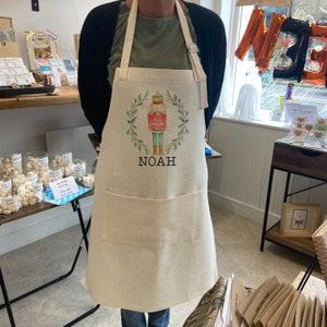 Puede incluir: Delantal beige con un dise&ntilde;o de cascanueces y el nombre "NOAH" impreso. El delantal presenta una corona verde y bayas rojas. Tiene un bolsillo frontal grande y una correa ajustable para el cuello. La tela es de color natural.