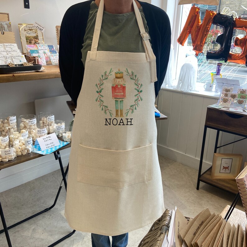 Puede incluir: Delantal beige con un dise&ntilde;o de cascanueces y el nombre "NOAH" impreso. El delantal presenta una corona verde y bayas rojas. Tiene un bolsillo frontal grande y una correa ajustable para el cuello. La tela es de color natural.