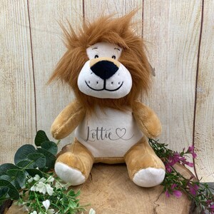 Può includere: Un leone di peluche marrone con un petto bianco, un naso nero e occhi neri. Il leone ha una criniera soffice ed è seduto su una superficie di legno. Il petto del leone è ricamato con il nome "Lottie" e un cuore.