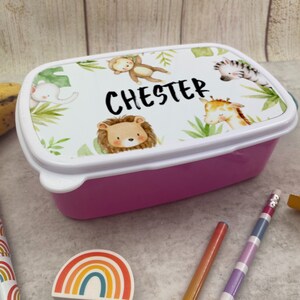 Puede incluir: Una fiambrera rectangular rosa con una tapa blanca con un dise&ntilde;o de animales de la jungla y el nombre "Chester". La tapa tiene un borde blanco y presenta un mono, un elefante, un le&oacute;n, una jirafa y una cebra.