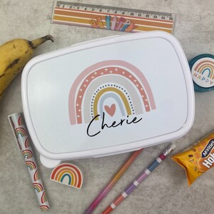 May include: White lunchbox with a rainbow design and the name "Cherie" in black script. The image includes a banana, pencils, a pen, a ruler, a sticker that says "Happy", and a snack.