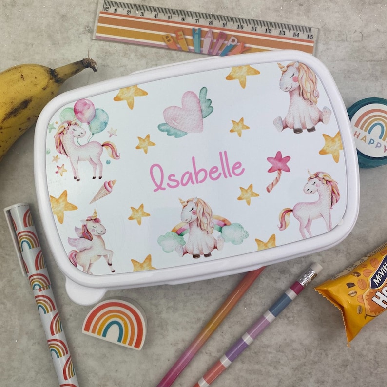 May include: White rectangular lunchbox decorated with unicorns, stars, and a heart with wings. The name "Isabelle" is written in pink script. A banana, pencils, and a rainbow eraser are also visible. Other stationery items are also present.