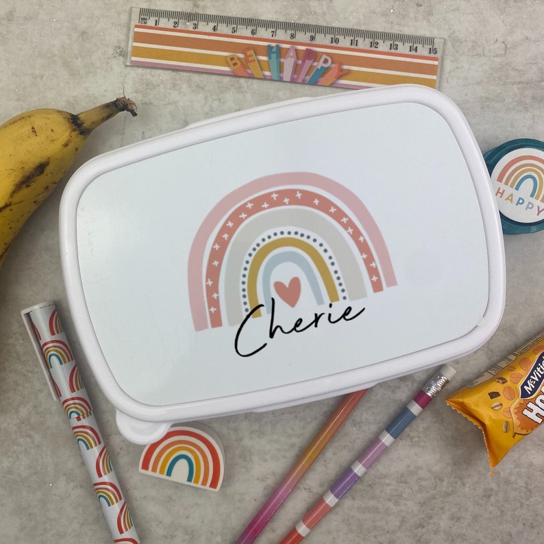 May include: White lunchbox with a rainbow design in pastel colors, a heart, and the name "Cherie" in script. A banana, pencils, a ruler, and other stationery items are scattered around the lunchbox.