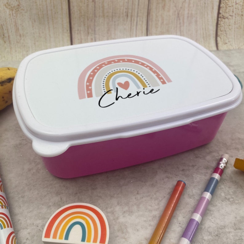 May include: A pink rectangular lunchbox with a white lid. The lid features a rainbow design with a heart and the name "Cherie" in black script. The lunchbox is surrounded by pencils and a rainbow eraser.