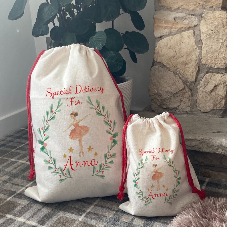 May include: Two off-white drawstring gift bags with red trim and drawstrings. Each bag features a watercolor ballerina design with the text "Special Delivery For Anna". The design includes a green wreath and gold stars.