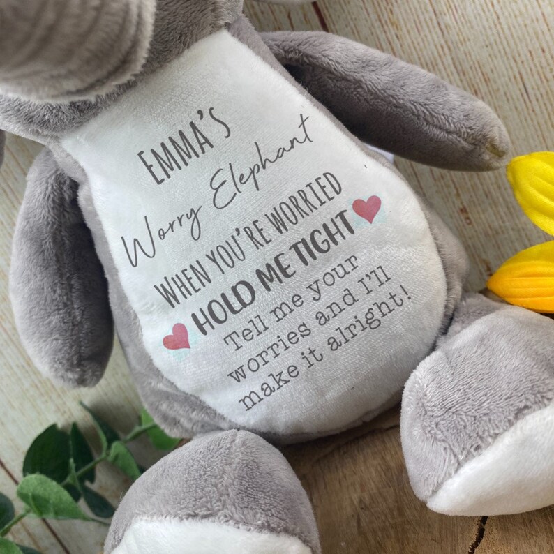 Op de afbeelding: Een grijze pluche olifant met een witte buikvlek. De vlek bevat de tekst "EMMA'S Worry Elephant" en een geruststellende boodschap. Twee kleine rode hartjes zijn op de vlek gedrukt. De olifant is een zacht, knuffelbaar speelgoed.