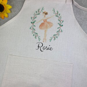 Puede incluir: Delantal beige claro con bolsillo y lazos. Presenta una ilustraci&oacute;n a la acuarela de una bailarina con tut&uacute; melocot&oacute;n, enmarcada por una corona verde con bayas rojas. El nombre "Rosie" est&aacute; escrito debajo en escritura negra.