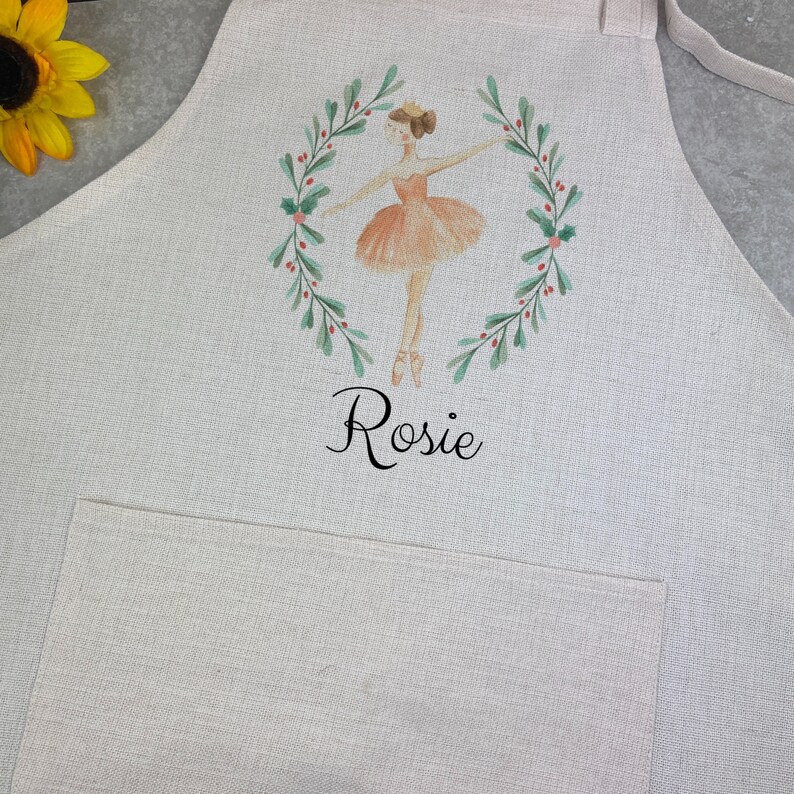 Puede incluir: Delantal beige claro con bolsillo y lazos. Presenta una ilustraci&oacute;n a la acuarela de una bailarina con tut&uacute; melocot&oacute;n, enmarcada por una corona verde con bayas rojas. El nombre "Rosie" est&aacute; escrito debajo en escritura negra.